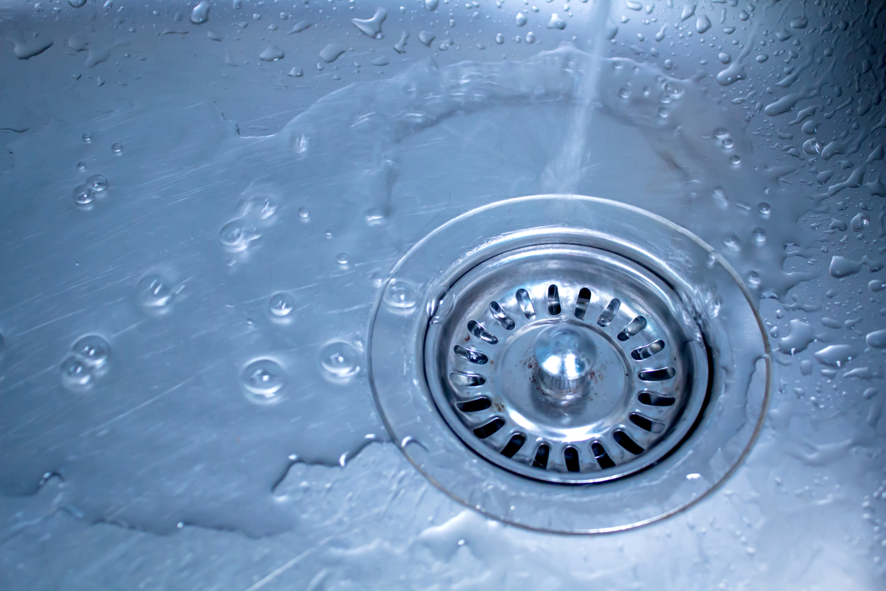 Close-up of water flowing into a kitchen sink drain, with droplets on the metal surface and a clear view of the drain strainer.