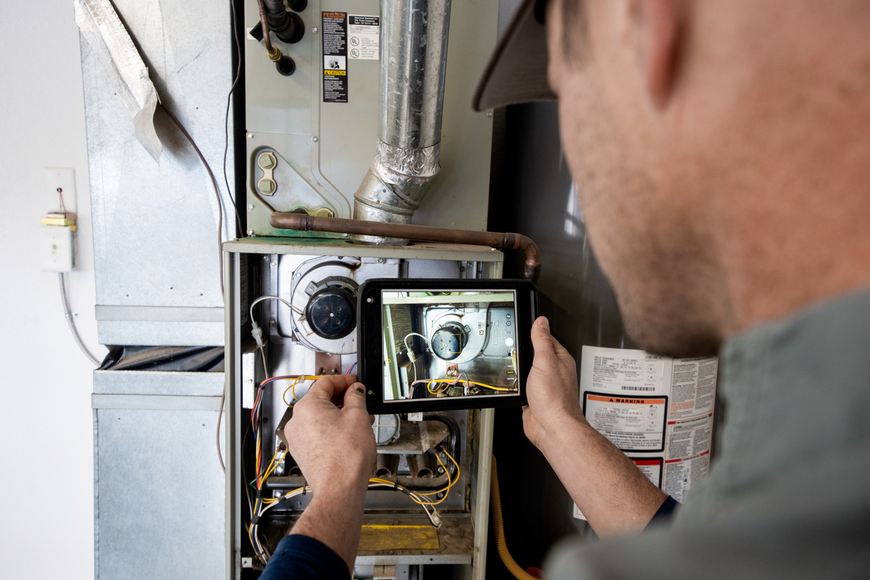 HVAC technician inspects the inside of a furnace with the front panel open, holding a tablet that shows a close-up view of the furnace components and wiring.