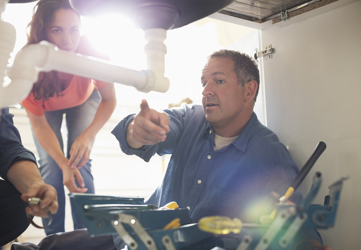 A plumber explains the plumbing under a sink to a woman, emphasizing the importance of inspecting home pipes.