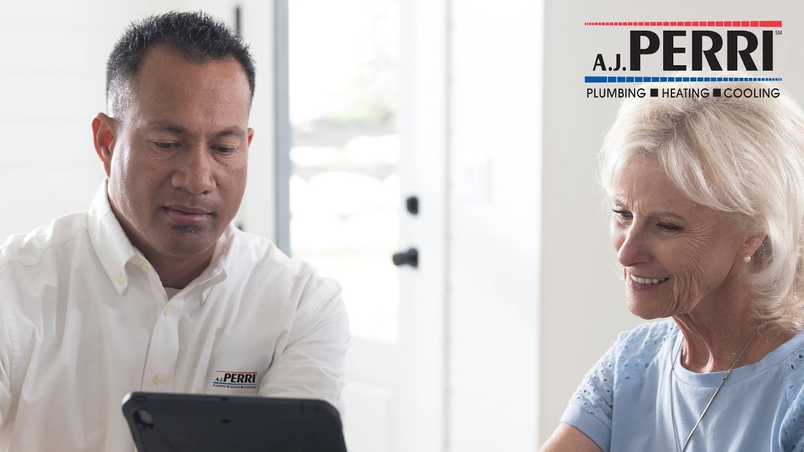 A technician from A.J. Perri Plumbing, Heating, and Cooling shows information on a tablet to a smiling older woman in her home.
