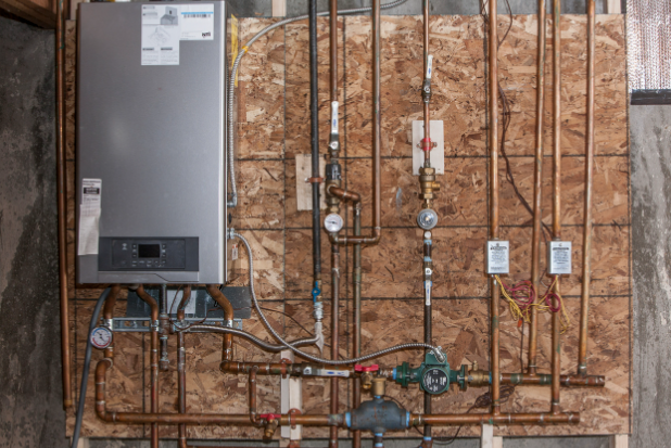 Close-up of a hydronic boiler installation on a wall, featuring a wall-mounted unit, copper pipes, pressure gauges, and various valves and controls.
