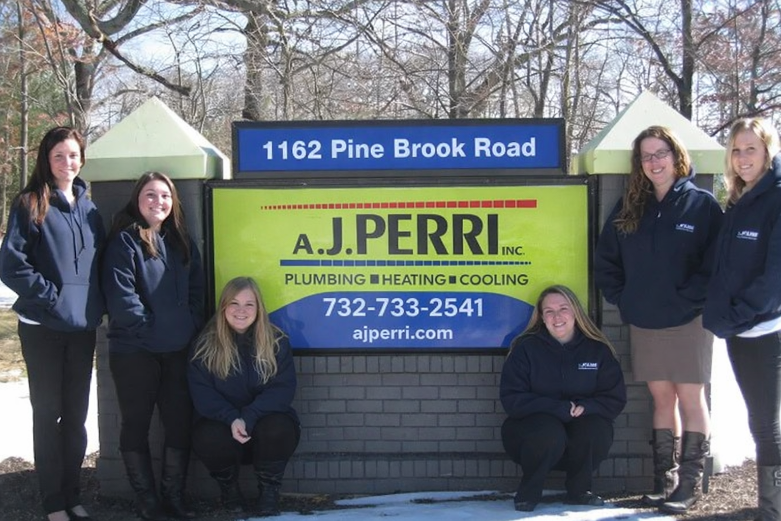 Six A.J. Perri employees pose in front of a roadside company sign at 1162 Pine Brook Road, which displays the phone number 732-733-2541 and the website ajperri.com.